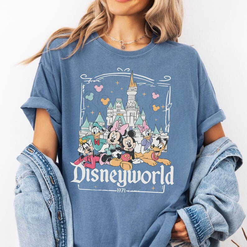 May include: Blue t-shirt featuring a graphic of Disney characters in front of a castle. The word "Disneyworld" is printed in a white script font, with "1971" below. The shirt has rolled-up sleeves.