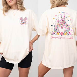 May include: Cream-colored t-shirt featuring a floral Mickey Mouse design on the front and a floral castle design on the back. The text "Happiest place on earth" is printed on both sides of the shirt. The shirt has short sleeves.
