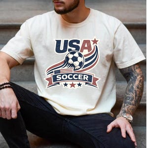 May include: A cream-colored short-sleeve t-shirt featuring a graphic design with the text USA SOCCER. The design includes a soccer ball, a small American flag, red and blue swooping lines, and stars in a vintage-inspired patriotic color scheme.