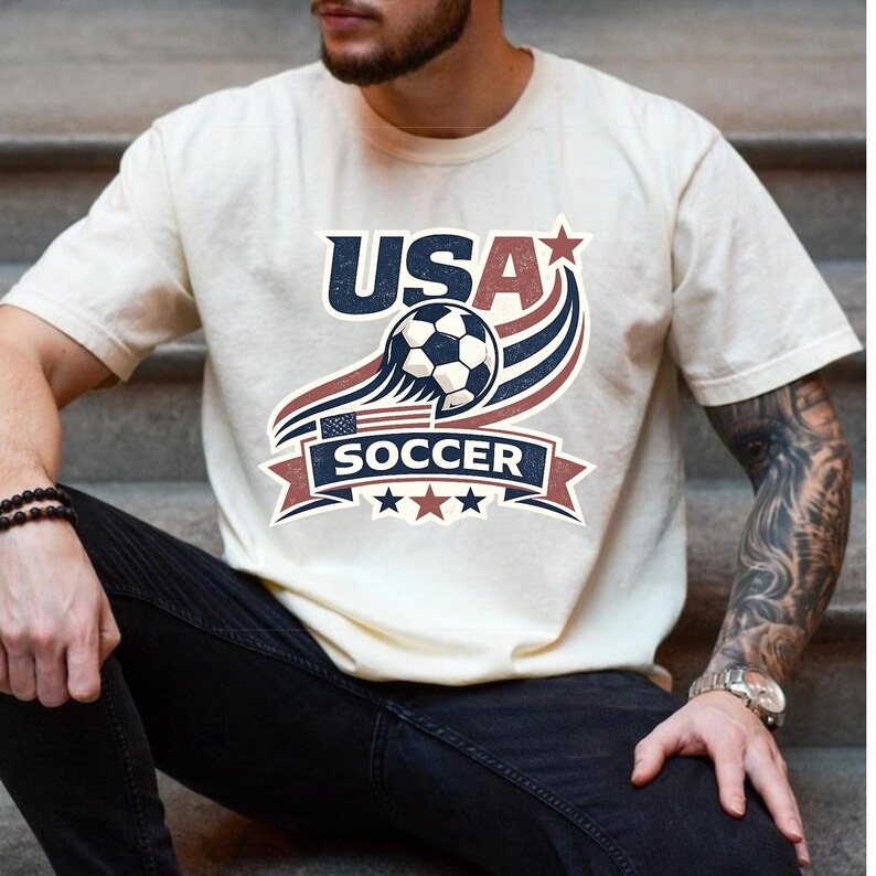 May include: A cream-colored short-sleeve t-shirt featuring a graphic design with the text USA SOCCER. The design includes a soccer ball, a small American flag, red and blue swooping lines, and stars in a vintage-inspired patriotic color scheme.