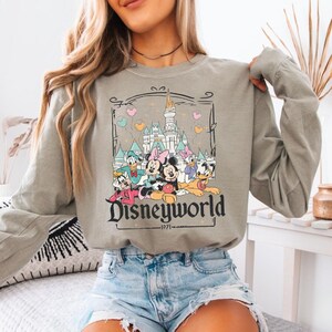 May include: A long-sleeved, olive green sweatshirt featuring a graphic of Disney characters including Mickey and Minnie Mouse, Donald Duck, and Daisy Duck. The graphic also includes a castle and the text "Disneyworld - 1971".