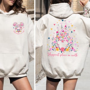 May include: Cream-colored hoodie featuring a floral Mickey Mouse design on the front and a floral castle design on the back. The text "Happiest place on earth" is printed on both sides. The designs incorporate pink, yellow, and blue flowers.