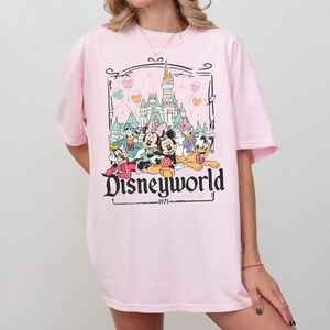 May include: A light pink t-shirt featuring a graphic of Disney characters in front of a castle. The graphic includes Mickey Mouse, Minnie Mouse, Donald Duck, Daisy Duck, and Goofy. The text "Disneyworld" and "1971" are also present.