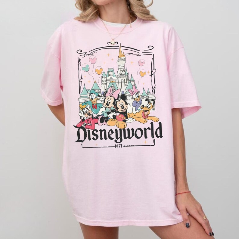 May include: A light pink t-shirt featuring a graphic of Disney characters in front of a castle. The graphic includes Mickey Mouse, Minnie Mouse, Donald Duck, Daisy Duck, and Goofy. The text "Disneyworld" and "1971" are also present.