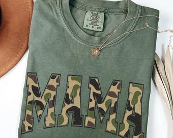 May include: An olive green t-shirt with the word "MAMA" in a camouflage pattern across the front. The shirt is folded and has a gold necklace with a pendant. The shirt is made by Comfort Colors.
