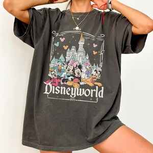 May include: Dark gray t-shirt featuring a graphic of Disney characters in front of a castle. The word "Disneyworld" is printed in white, along with the year "1971". The shirt has a vintage, worn look.