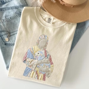 May include: Cream-colored t-shirt featuring an embroidered design of a figure holding a lamb. The figure's clothing is patchwork with floral and checkered patterns in pastel colors. The shirt is folded on top of blue jeans and a tan hat.