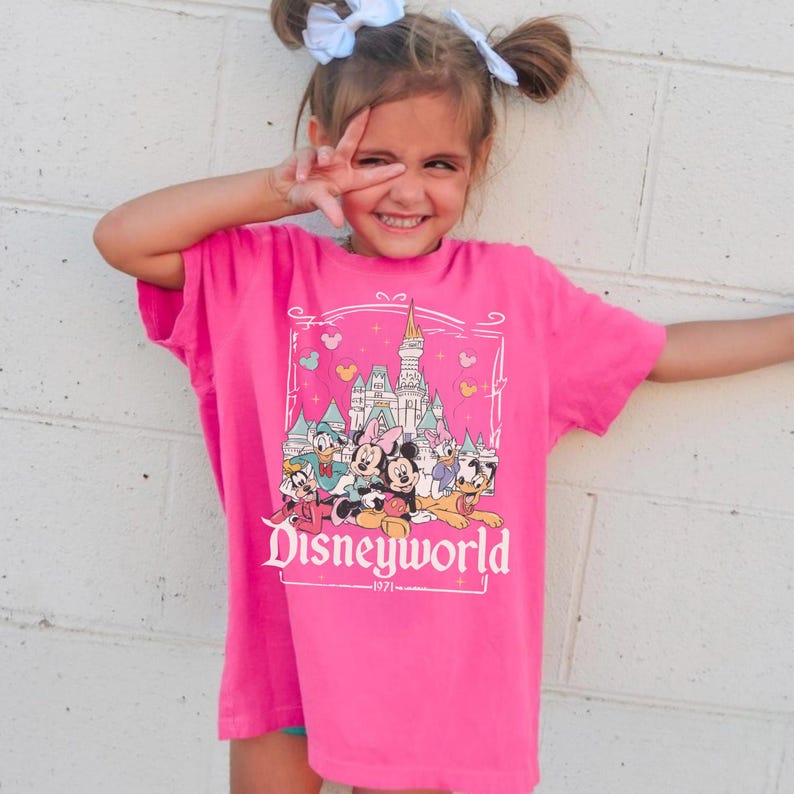 May include: A bright pink t-shirt featuring a Disney World graphic. The graphic includes a castle, Mickey and Minnie Mouse, Donald Duck, and other characters. The text "Disneyworld" is displayed prominently.