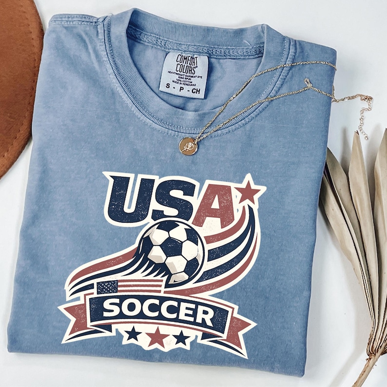 May include: A blue short-sleeved t-shirt featuring a graphic print with the text USA SOCCER, a soccer ball, a small American flag, and red and blue stars. The shirt has a Comfort Colors brand label on the inside neck and a crew neckline.
