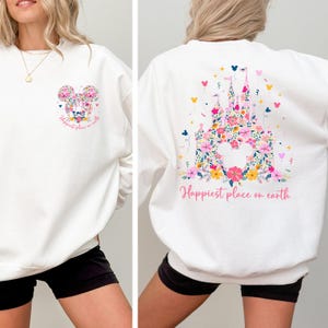 May include: White sweatshirt featuring a floral Mickey Mouse design on the front and a floral castle design on the back. The text "Happiest place on earth" is printed on both sides in a script font. The sweatshirt is a casual style.
