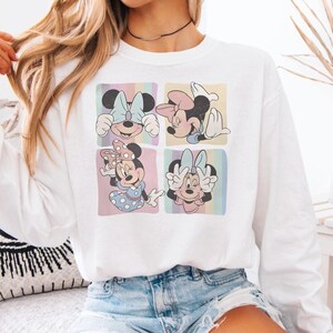 Puede incluir: Sudadera blanca con cuatro paneles que muestran ilustraciones de Minnie Mouse. Cada panel muestra una pose diferente de Minnie Mouse sobre un fondo pastel. La sudadera es de cuello redondo y manga larga.