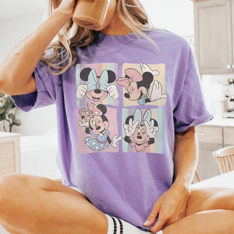 Puede incluir: Una camiseta lila con cuatro paneles cuadrados con ilustraciones de Minnie Mouse. Cada panel muestra una pose diferente de Minnie Mouse, con rayas de arco&iacute;ris pastel de fondo. La camiseta tiene un corte holgado y extragrande.