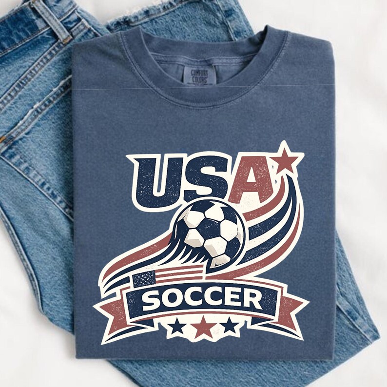 May include: A blue t-shirt featuring a graphic with the text USA SOCCER, a soccer ball, and stylized red, white, and blue stripes. The design includes a small American flag and three stars at the bottom, all presented in a distressed, vintage-inspired print style.