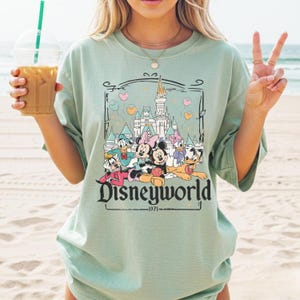 May include: A light green t-shirt featuring a Disneyworld graphic with Mickey Mouse, Minnie Mouse, Donald Duck, Daisy Duck, and Goofy. The graphic includes a castle and the text "Disneyworld - 1971".