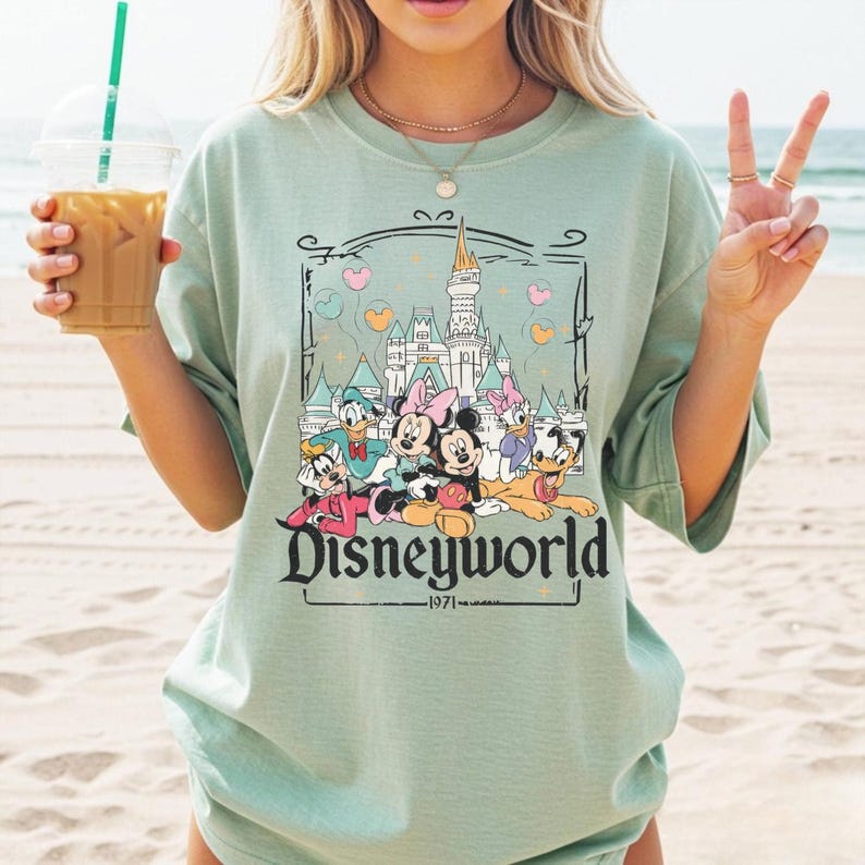 May include: A light green t-shirt featuring a Disneyworld graphic with Mickey Mouse, Minnie Mouse, Donald Duck, Daisy Duck, and Goofy. The graphic includes a castle and the text "Disneyworld - 1971".