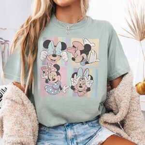 Puede incluir: Una camiseta verde claro con un gr&aacute;fico colorido de Minnie Mouse en cuatro poses diferentes. Cada pose est&aacute; sobre un fondo pastel. La camiseta es de estilo c&oacute;modo e informal.