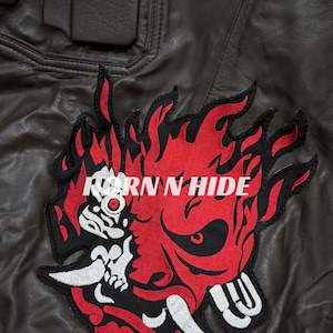 Pu&ograve; includere: Giacca di pelle marrone con una grande toppa ricamata con un disegno di demone fiammeggiante rosso e nero con la scritta "HORN N HIDE". La parola "SAMURAI" &egrave; visibile in alto a destra.