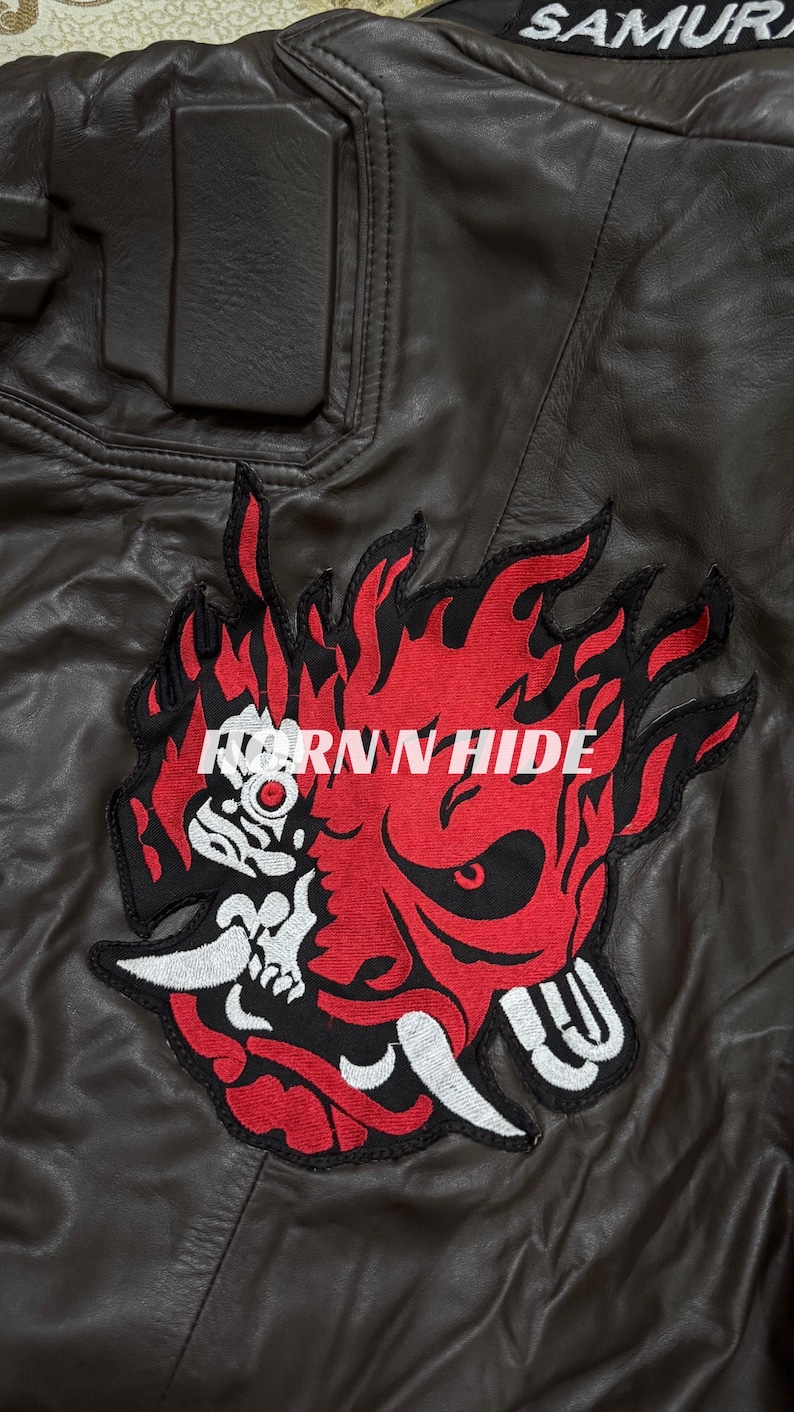 Pu&ograve; includere: Giacca di pelle marrone con una grande toppa ricamata con un disegno di demone fiammeggiante rosso e nero con la scritta "HORN N HIDE". La parola "SAMURAI" &egrave; visibile in alto a destra.