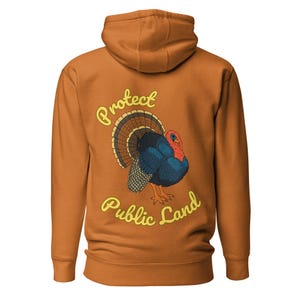Könnte beinhalten: Ein orangefarbener Kapuzenpullover mit Kapuze und langen Ärmeln. Auf der Rückseite befindet sich eine farbenfrohe Illustration eines Truthahns mit dem Text "Protect Public Land" in gelber, kursiver Schrift. Der Truthahn hat blaue, rote und braune Federn.