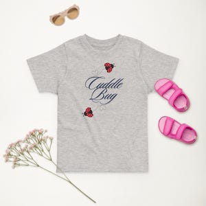Puede incluir: Una camiseta gris claro con las palabras "Cuddle Bug" en escritura azul marino, adornada con gráficos de mariquitas rojas y negras. También hay sandalias y gafas de sol rosas.