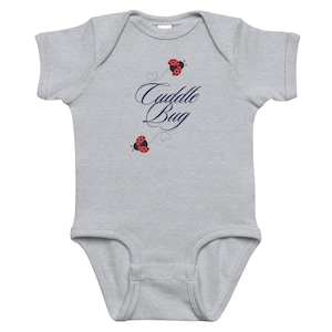 Puede incluir: Un body de bebé gris claro con mangas cortas. El body presenta las palabras "Cuddle Bug" en escritura azul marino, junto con dos ilustraciones de mariquitas rojas. Las mariquitas tienen lunares negros y están situadas encima y debajo del texto.