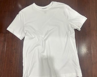 Może przedstawiać: Zwykły biały t-shirt z krótkim rękawem. Koszulka ma okrągły dekolt i jest wykonana z miękkiej, lekkiej tkaniny. Koszulka leży na ciemnobrązowej drewnianej powierzchni.