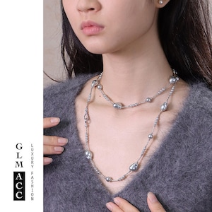 Op de afbeelding: Een zilveren ketting met twee strengen, met grote, onregelmatig gevormde grijze parels en kleinere zilveren kralen. De ketting wordt gedragen over een grijze trui. De afbeelding bevat ook de tekst "GLM ACC" en "LUXURY FASHION".