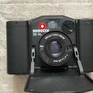 Op de afbeelding: Zwarte Minox 35 AL camera met een rood lampje. De camera heeft een lens met de tekst "Minox Color-Minar 1:4 = 35 mm" en een ISO 100-instelling. De lensklep van de camera is open.