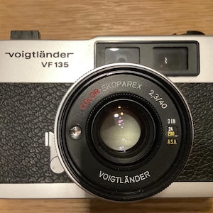 Könnte beinhalten: Eine silber-schwarze Voigtländer VF 135 Filmkamera mit einem Color-Skoparex 2,3/40 Objektiv. Das Gehäuse zeigt den Markennamen und das Modell, einen strukturierten schwarzen Griff, ein zentrales Objektiv, einen Sucher und Filmempfindlichkeit.
