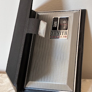 May include: A vintage Zenith Royal 16 transistor radio encased in a black leather-like folding cover. The radio features a silver-toned metal speaker grille, a black tuning dial with white numbers 9, 11, and 16, and a small crown logo emblem.