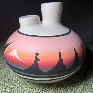 May include: A matte ceramic vessel with two spouts, featuring a hand-painted sunset landscape. The design displays a gradient of pink and orange hues, black silhouettes of pine trees, and a white-capped mountain peak against a dark horizon.