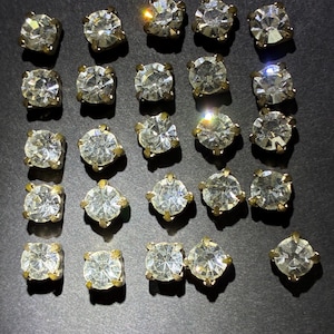 May include: A collection of twenty-one round, clear faceted rhinestones set in gold-toned metal prong settings. Each individual piece features a circular cut stone with multiple reflective facets, arranged in a grid-like pattern on a dark surface.