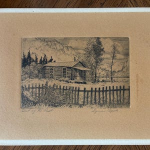 May include: A black and white ink drawing of a log cabin set behind a wooden picket fence, with trees and mountains in the background. The artwork is titled Out of the Past and signed by Lyman Byxbe, mounted on a tan mat with a white border.
