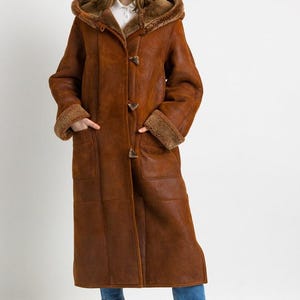 May include: A long, brown, hooded coat with a textured exterior and a plush, tan-coloured faux fur lining visible at the hood, cuffs, and front opening. The coat features three toggle closures down the centre and two large patch pockets on the front.