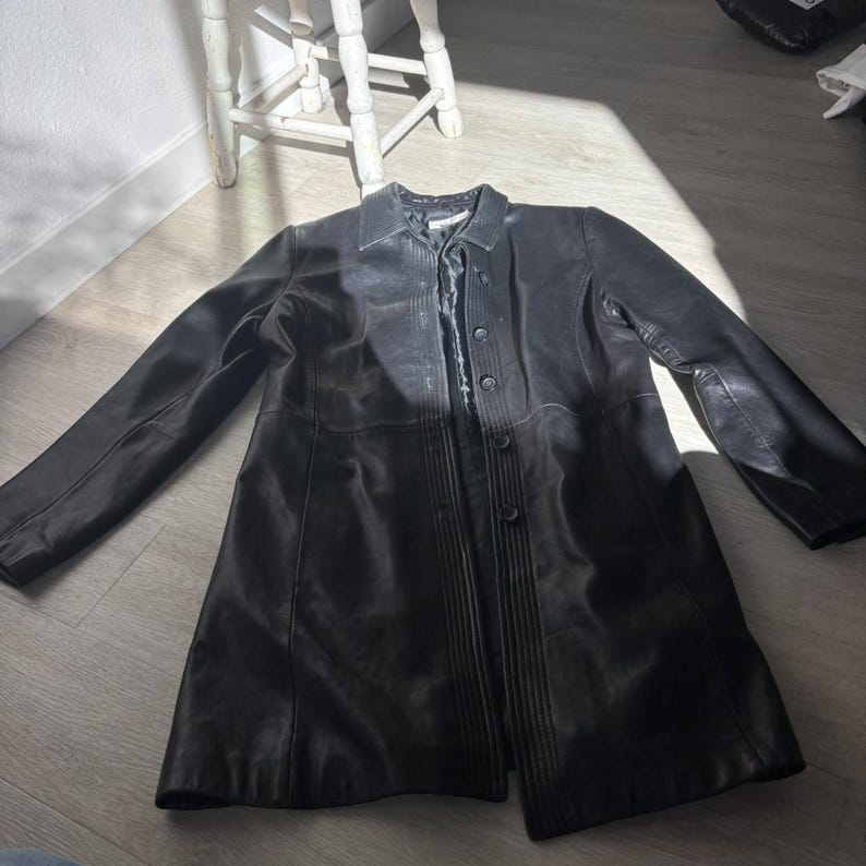 May include: A black leather coat with a button-up front and a collared neckline. The coat is long, reaching to the mid-thigh, and has a classic, tailored design. The coat is lying flat on a wooden floor.