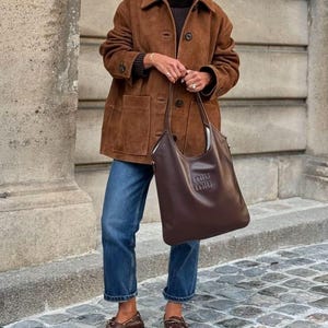 May include: A brown suede jacket with a collared neckline and button closure. The jacket is paired with blue jeans, brown leather loafers, and a brown leather tote bag with a logo. The outfit is completed with a dark brown turtleneck.