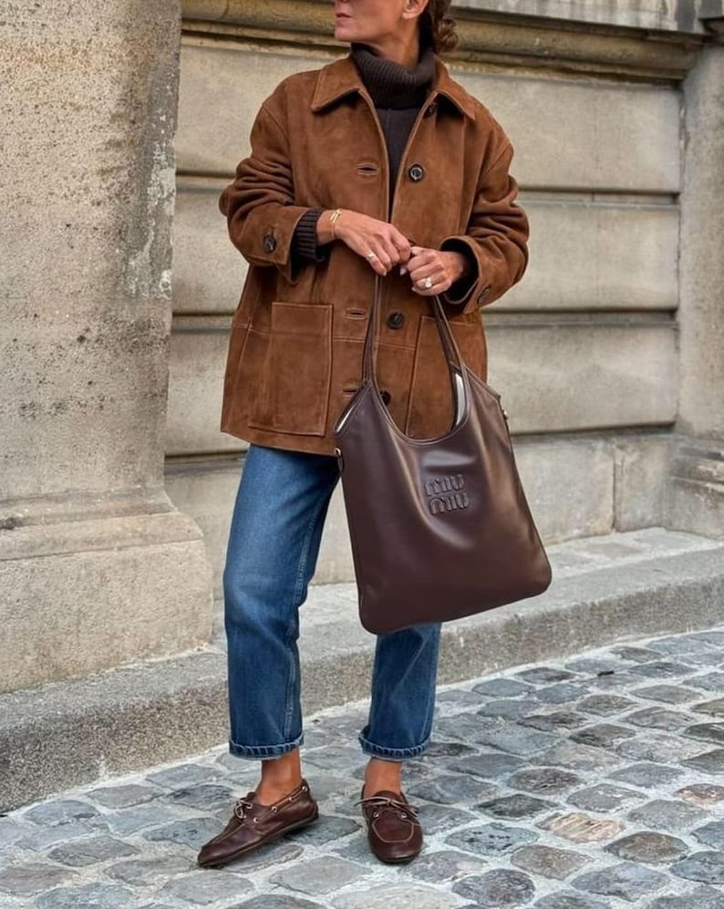May include: A brown suede jacket with a collared neckline and button closure. The jacket is paired with blue jeans, brown leather loafers, and a brown leather tote bag with a logo. The outfit is completed with a dark brown turtleneck.