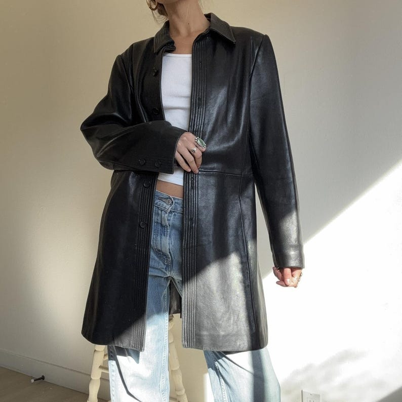 May include: A black leather coat with a button-up front and a collared neckline. The coat has long sleeves and a knee-length hem. The coat is worn over a white top and blue jeans. The coat is a classic and versatile piece of outerwear.