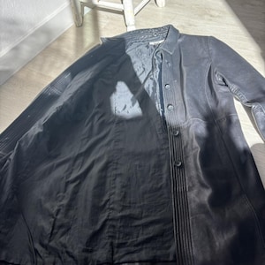 May include: A long, black coat with a button-up front and a collared neckline. The coat appears to be made of a soft material. The coat is lying on a light-coloured wooden floor.