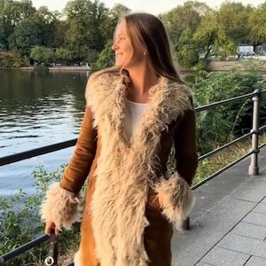 May include: A long brown coat with cream-colored faux fur trim. The coat features a wide collar and cuffs, and is worn over a white top and blue jeans. A dark brown handbag is held in the hand.