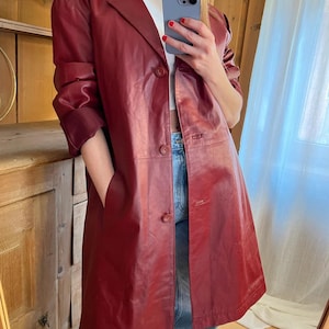 May include: A burgundy leather coat with a classic lapel collar and three button closure. The coat has long sleeves and pockets. The person is wearing a white top and blue jeans. The coat appears to be knee-length.