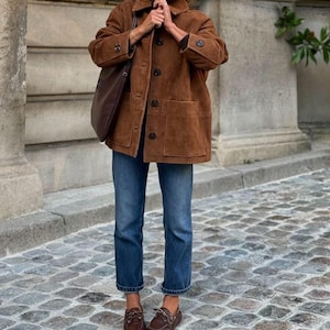 May include: A brown suede jacket with large pockets and dark buttons. The jacket is paired with blue jeans and brown leather boat shoes. A brown leather tote bag is carried over the shoulder.