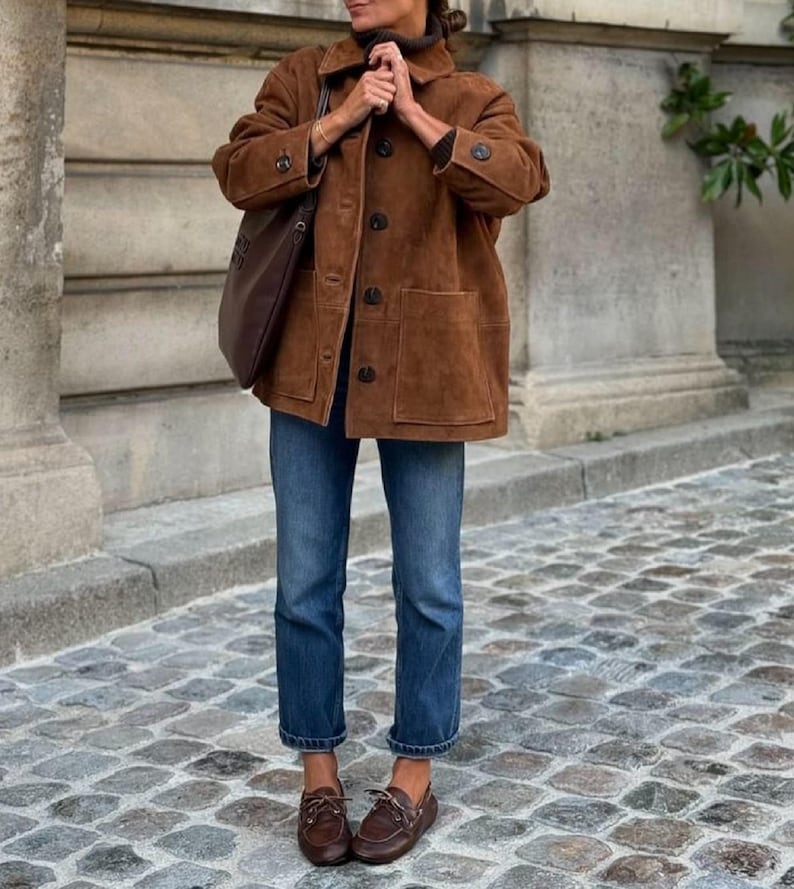 May include: A brown suede jacket with large pockets and dark buttons. The jacket is paired with blue jeans and brown leather boat shoes. A brown leather tote bag is carried over the shoulder.