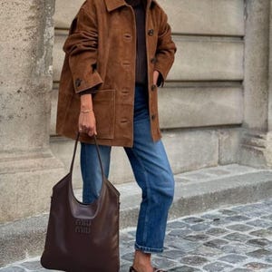 May include: A brown suede jacket with a button-up front, worn over a dark sweater and blue jeans. A large brown leather tote bag with the brand name embossed on the front is held in the hand. Brown leather boat shoes complete the outfit.