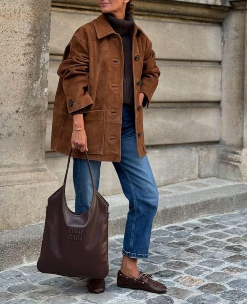 May include: A brown suede jacket with a button-up front, worn over a dark sweater and blue jeans. A large brown leather tote bag with the brand name embossed on the front is held in the hand. Brown leather boat shoes complete the outfit.