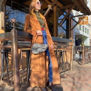 Puede incluir: Un abrigo largo de ante marrón con un corte entallado. El abrigo se lleva con vaqueros azules, botas marrones y un bolso estampado. El conjunto se complementa con gafas de sol y un collar. La longitud del abrigo llega hasta los tobillos.