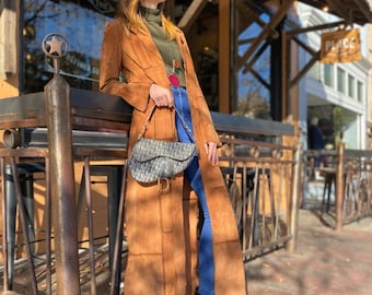 May include: A long, brown suede coat with a tailored fit. The coat is worn with blue jeans, brown boots, and a patterned handbag. The outfit is accessorized with sunglasses and a necklace. The coat's length extends to the ankles.