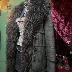 May include: A dark green coat with a long, shaggy, dark brown fur collar and trim. The coat has toggle closures and pockets. A mannequin head is wearing a silver cross pendant with a purple stone.