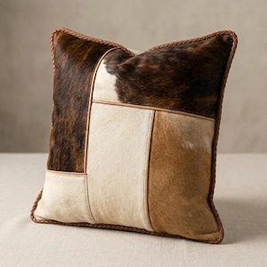 May include: Square decorative pillow with a patchwork design. The pillow features sections of brown and white cowhide, with a braided brown border. The pillow has a natural, rustic aesthetic.