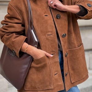 May include: A brown suede jacket with large pockets and dark buttons. A matching brown leather tote bag with a white interior is visible. The jacket is paired with blue jeans. A gold bracelet is worn on the wrist.